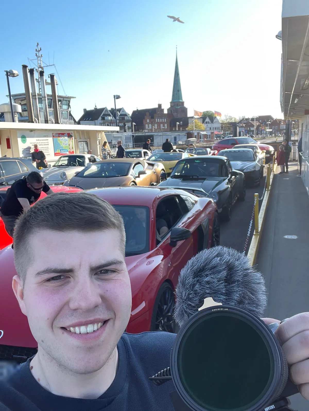 Selfie mit Kamera vor den geparkten Sportwagen, im Hintergrund ein Kirchturm